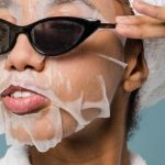 Routines - Self esteem young African American lady with moisturizing sheet mask and towel on hand wearing trendy sunglasses while standing against blue background in bathrobe after shower